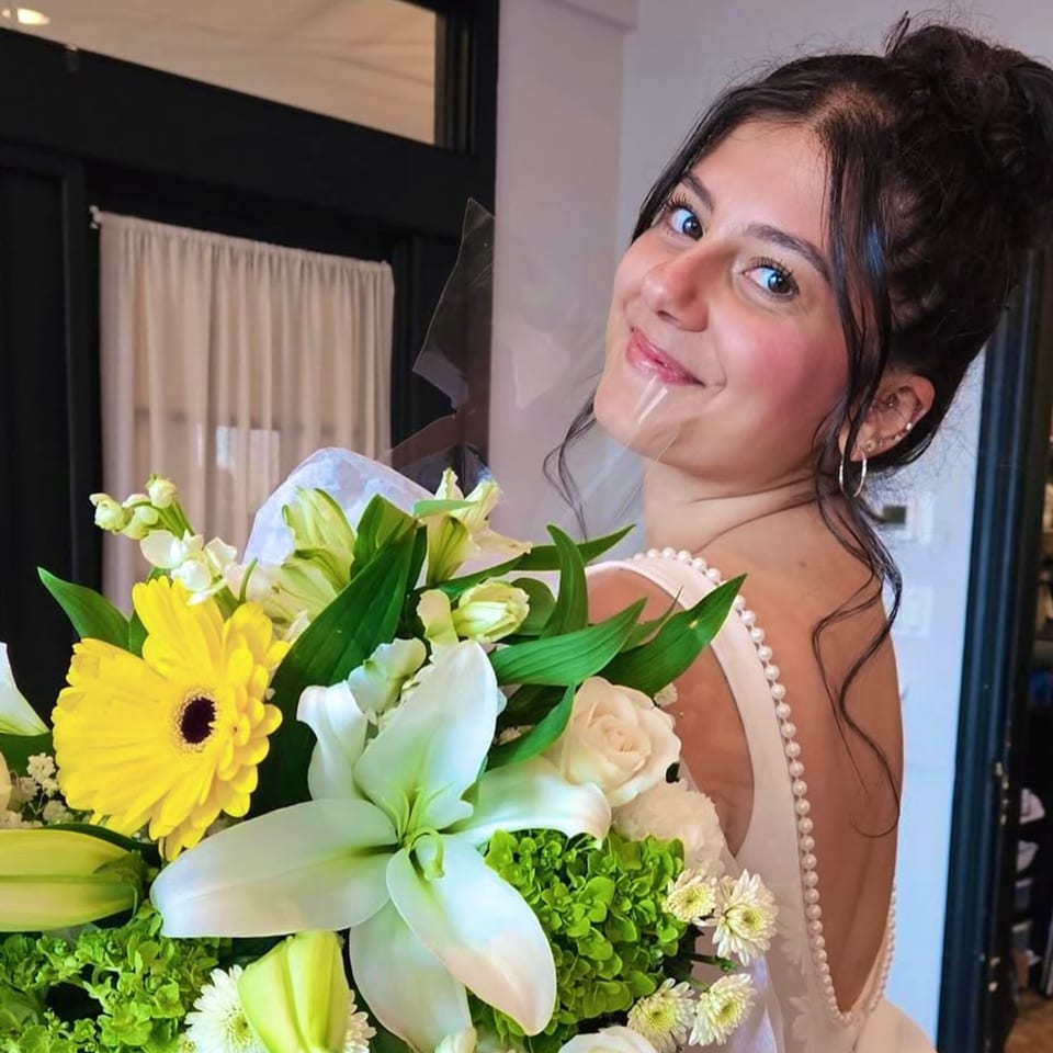 Smiling woman in a pearl-trimmed dress holding a vibrant bouquet of yellow, white, and green flowers.