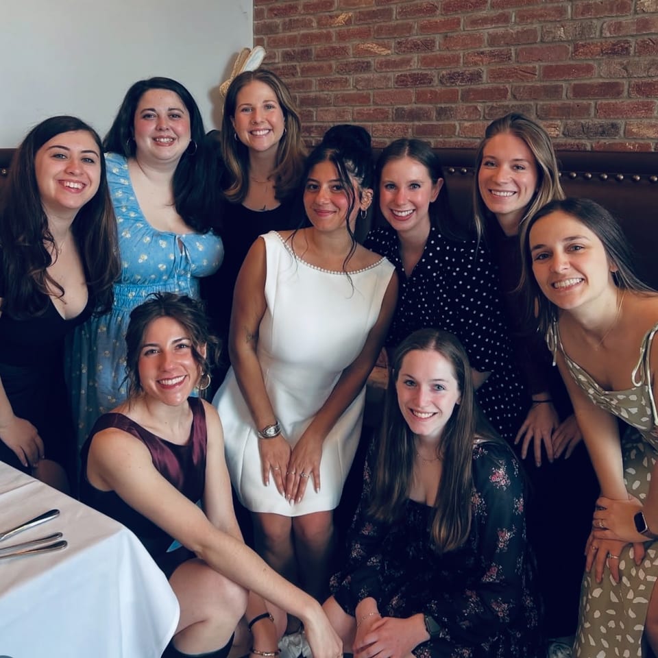 Nine smiling women pose together in a restaurant with a brick wall and booth seating.