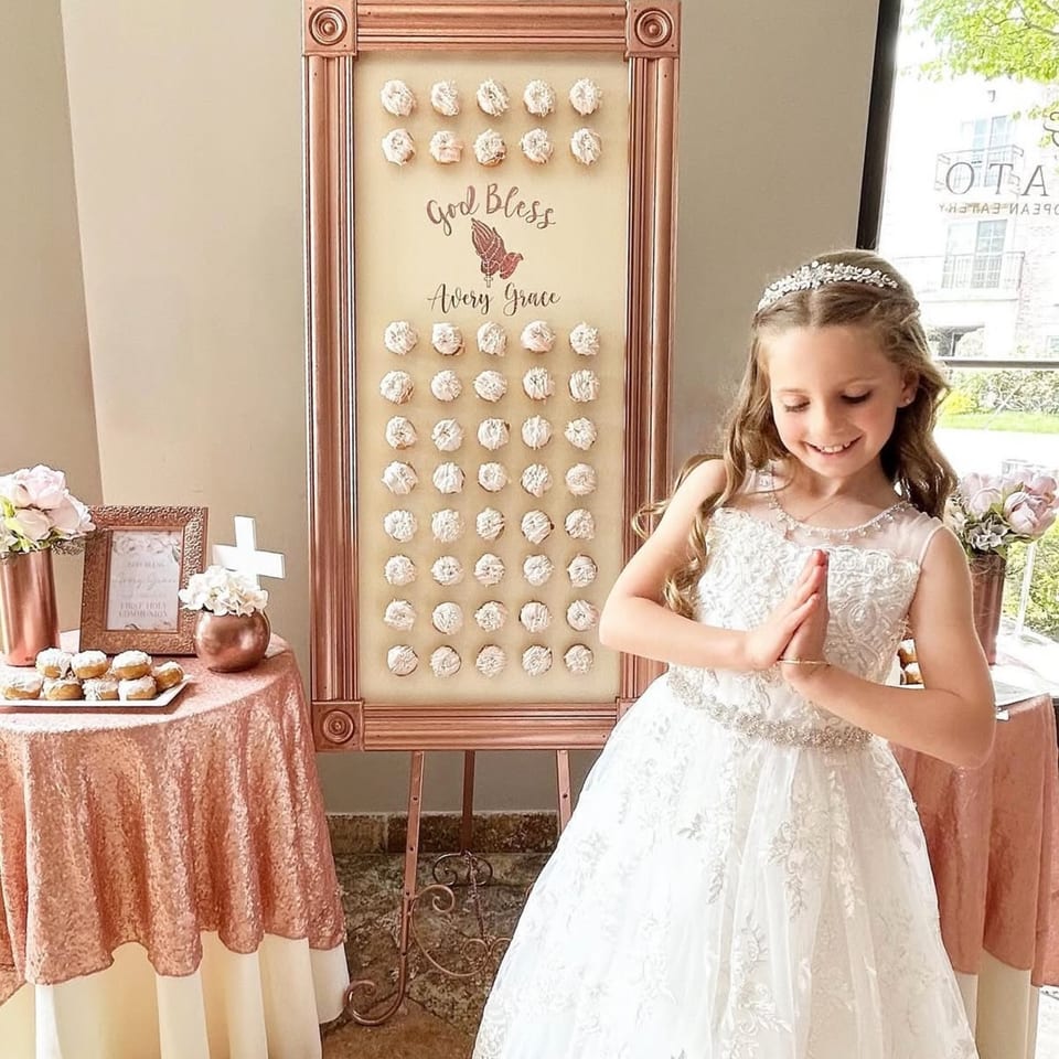Young girl in white dress praying beside a "God Bless Avery Grace" sign with cupcakes.