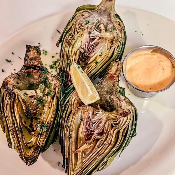 Three grilled artichoke halves with a lemon wedge and a side of dipping sauce on a white plate.