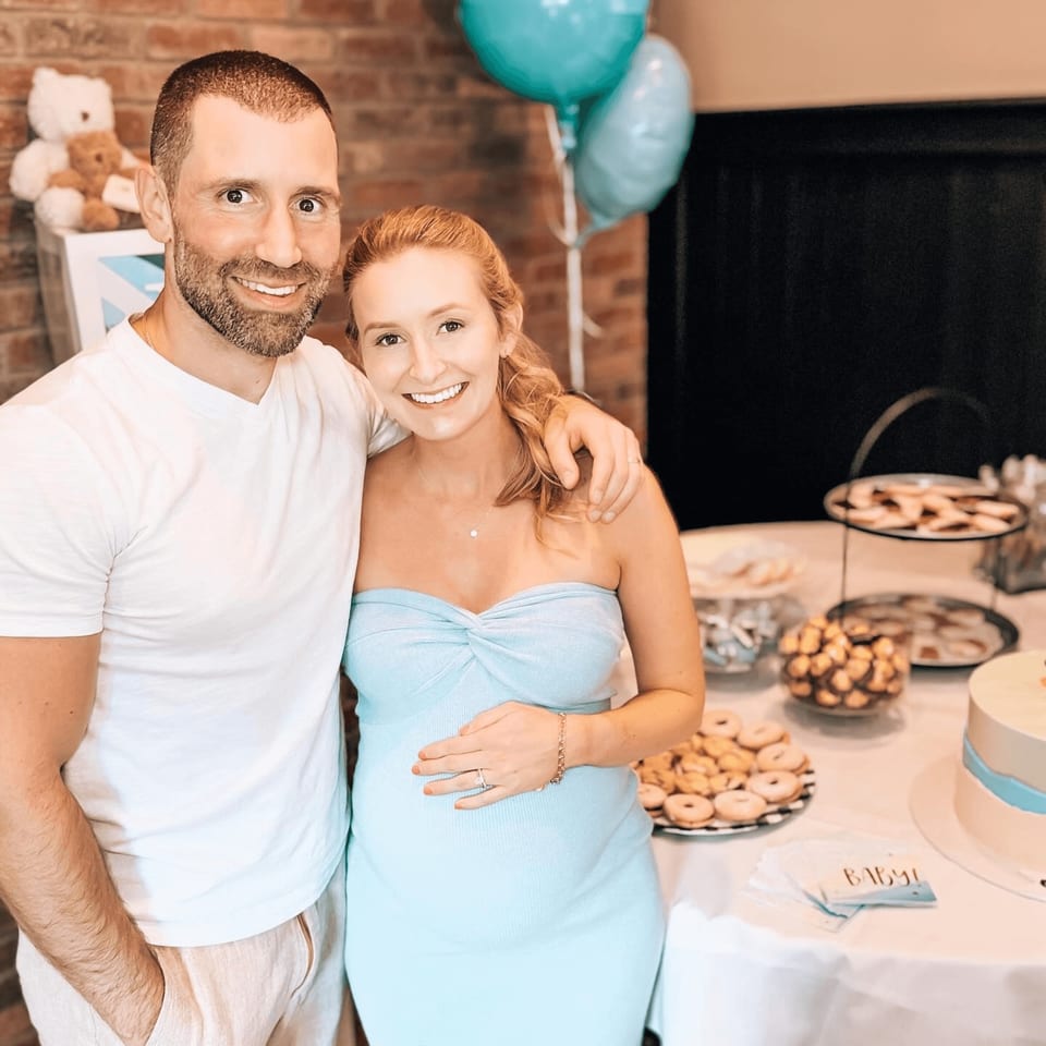 A smiling couple at a baby shower, with the pregnant woman in a light blue dress.