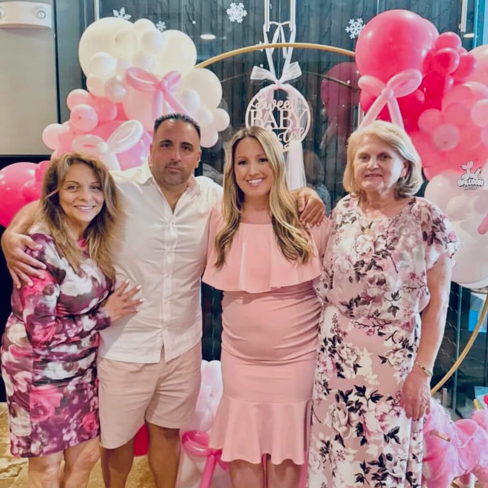 Four people, two men and two women, pose in front of pink and white balloons at a baby shower.