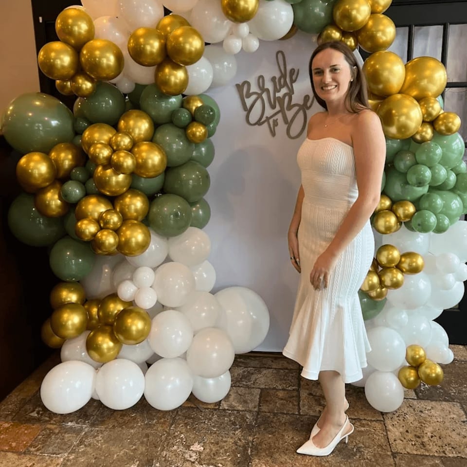 A smiling woman in a white dress stands before a "Bride to Be" sign and balloon arch.
