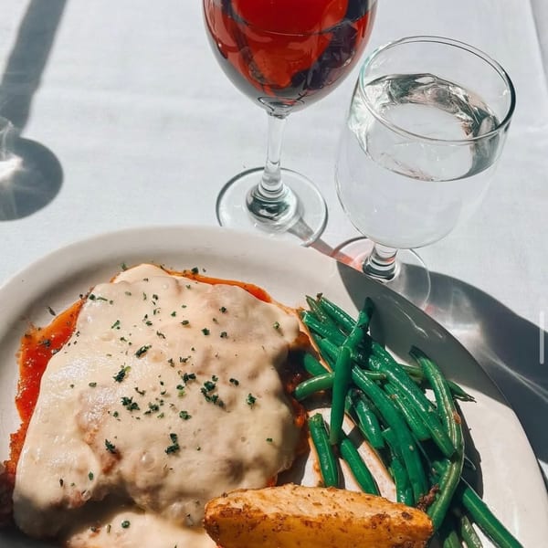 Chicken parmesan with green beans, a potato wedge, and two glasses on a white tablecloth.