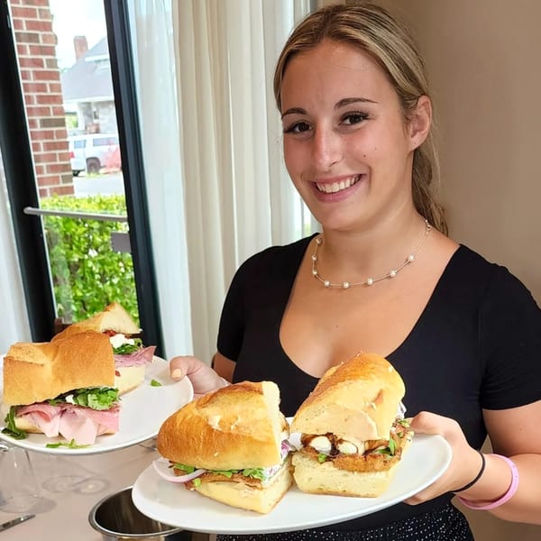 A smiling server holds two plates with large Italian sandwiches, one with ham and arugula.