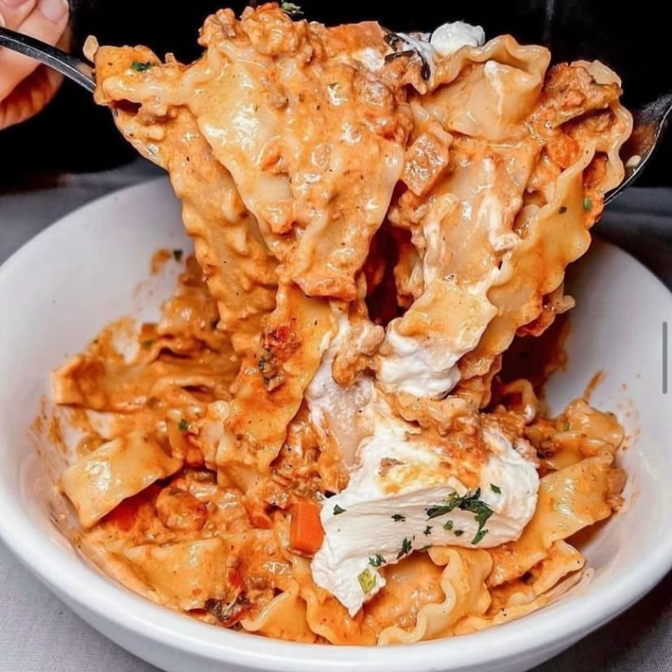 A fork lifts creamy, ruffled pasta with meat sauce and dollops of ricotta from a white bowl.