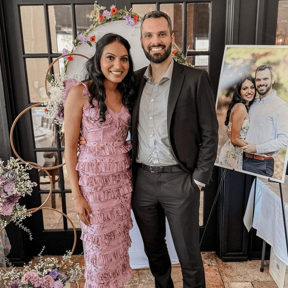 Smiling couple posing in front of a floral decorated archway and a large framed photo.