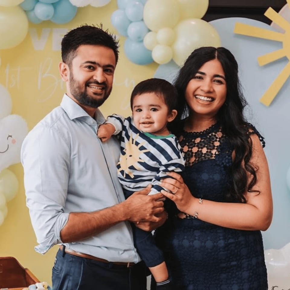 A smiling family, a man, woman, and baby, pose in front of a balloon arch.