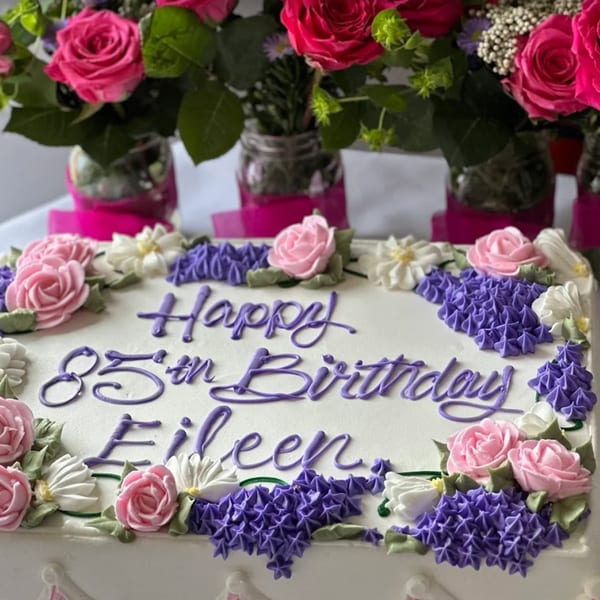 White birthday cake with purple "Happy 85th Birthday Eileen" text and pink and purple frosting flowers.